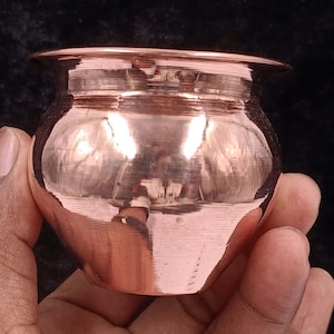 May include: A small, copper-coloured pot with a rounded body and a flared rim. The pot is held in a hand, and the polished surface reflects the surrounding environment. The pot's design is simple and traditional, suggesting a possible use for cooking or storage.