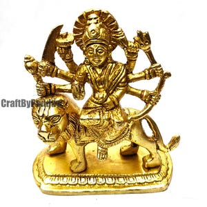 May include: A golden brass statue of a multi-armed deity riding a lion. The statue has intricate details, including the deity's jewelry and the lion's mane. The base is rectangular and the overall design is ornate.