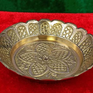 May include: A brass bowl with a detailed floral pattern in the center. The bowl has a scalloped edge and a gold-coloured interior. The bowl is set against a red background. Likely used for decorative purposes.