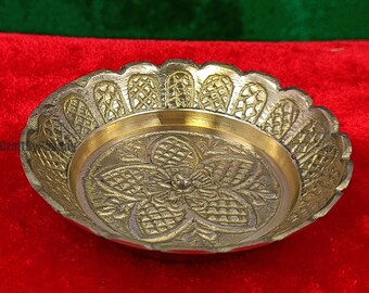 May include: A decorative brass bowl with an intricate floral design in the center. The bowl has a scalloped edge and a gold-colored interior, set against a red background. The bowl is likely used for decorative purposes.