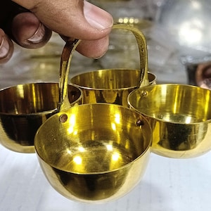 Op de afbeelding: Een goudkleurige metalen serveerschaal met vier kleine kommen en een handvat. De kommen zijn rond en hebben een glanzende afwerking. Het handvat is gebogen en verbindt de kommen voor gemakkelijk dragen. De schaal wordt waarschijnlijk gebruikt voor het serveren van dips, sauzen of kleine porties.