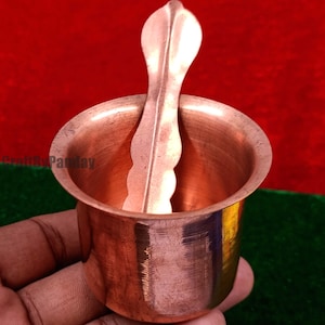 Puede incluir: Una taza de metal color cobre con una cuchara a juego. La taza tiene una base redondeada y un borde acampanado. La cuchara tiene un mango decorativo. Los artículos se sostienen en una mano sobre un fondo rojo y verde.