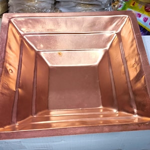 Può includere: Un bacino quadrato in metallo color rame con un design interno a gradini. Il bacino ha una superficie lucida e riflettente e sembra fatto a mano. I bordi sono leggermente arrotondati e la forma generale è simmetrica.