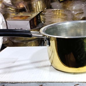 May include: A small, shiny gold-colored saucepan with a black handle. The interior of the pan is silver. The handle is attached with silver rivets. The pan is sitting on a white surface. The text "CraftByPanday" is visible on the left side.