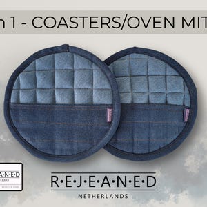 Può includere: Due sottobicchieri o presine rotonde in denim blu. La sezione superiore ha un design trapuntato e la sezione inferiore è in denim tinta unita. Il testo "2 in 1 - COASTERS/OVEN MITTS" è in alto. Il marchio "R.E.J.E.A.N.E.D NETHERLANDS" è in basso.