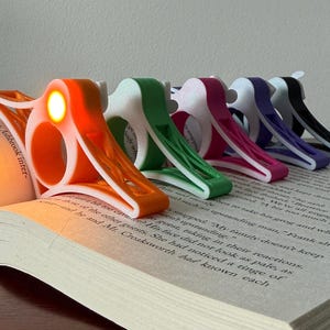 May include: A collection of colorful book lights, each clipped onto the pages of an open book. The lights are in various colors, including orange, green, pink, purple, and black. The orange light is illuminated.