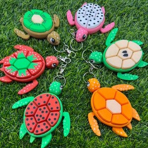 Op de afbeelding: Vijf kleurrijke schildpad-sleutelhangers met verschillende patronen. De sleutelhangers zijn gemaakt van plastic en hebben een glanzende afwerking. De patronen omvatten een kiwi, een watermeloen, een aardbei, een ananas en een effen ontwerp.