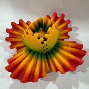 Op de afbeelding: Een 3D-geprinte bloemensculptuur met een levendige kleurovergang. De bloemblaadjes lopen over van rood naar geel en oranje, rondom een geel en groen centrum. Het ontwerp heeft ingewikkelde, gelaagde details.