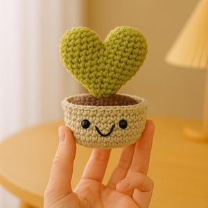 May include: A crocheted heart-shaped plant in a beige pot with a smiling face. The plant is green and the pot has black eyes and a black smile.