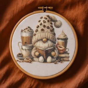 Puede incluir: Un bastidor de bordado muestra un gnomo leyendo un libro, con un gorro adornado con granos de café. La escena incluye tazas de café, libros y rosquillas, todo en tonos marrones cálidos.