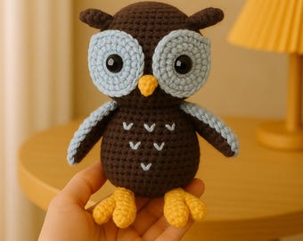 May include: A brown crocheted owl with blue eyes and yellow feet. The owl is sitting on a hand.