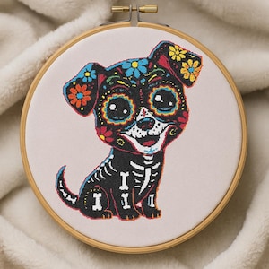 May include: A cross-stitch embroidery of a dog in a wooden hoop. The dog is decorated in a Day of the Dead sugar skull style, with floral and skeletal details in vibrant colors. The background is a light beige fabric.