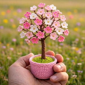 Peut inclure: Un petit arbre décoratif réalisé en crochet, doté d'un tronc marron et de nombreuses fleurs roses et blanches. L'arbre est planté dans un pot rond en crochet rose clair avec une base verte texturée représentant de la terre ou de l'herbe.