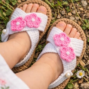 May include: Handmade white crochet sandals with pink flower accents and pearl embellishments. The sandals have a brown sole and are designed for a child. The sandals are shown on a grassy surface.