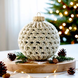 May include: A crocheted beige Christmas bauble in the shape of a sphere with a gold top. The bauble is sitting on a wooden surface with pine branches and pine cones.