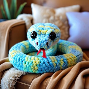 Pode incluir: Um brinquedo de pelúcia de cobra de crochê azul e amarelo com olhos vermelhos e língua vermelha. A cobra está enrolada em um sofá marrom.