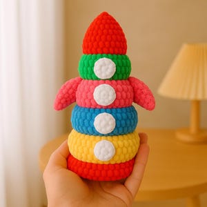 Könnte beinhalten: Ein farbenfrohes, gehäkeltes Stapelspielzeug in Form einer Rakete. Das Spielzeug besteht aus fünf Ringen in Rot, Grün, Rosa, Blau und Gelb. Jeder Ring hat einen weißen Kreis in der Mitte.