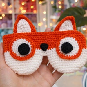 Puede incluir: Un antifaz para dormir tejido a mano con forma de cara de zorro. La pieza está hecha con hilo de color naranja brillante y blanco, presenta dos orejas puntiagudas, dos grandes ojos circulares con pupilas negras y bordes naranjas, y una pequeña nariz negra.