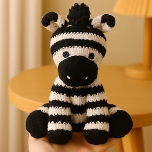 May include: A handmade, knitted zebra plush toy with black and white stripes. The zebra has a black snout, button eyes, and a small black pom-pom on its head. The toy is sitting on a light brown surface.