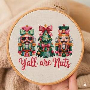 Peut inclure: Un cercle de broderie avec un motif de Noël. Le design comprend deux casse-noisettes et un sapin de Noël décoré. Le texte "Y'all are Nuts" est brodé en rouge. Le cercle est en bois.