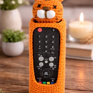 Pode incluir: Capa de controle remoto de crochê laranja com um design de rosto de tigre. A capa se encaixa em um controle remoto padrão, com recortes para os botões. O rosto do tigre tem detalhes brancos e detalhes costurados em preto. O controle remoto tem os números 1-9.