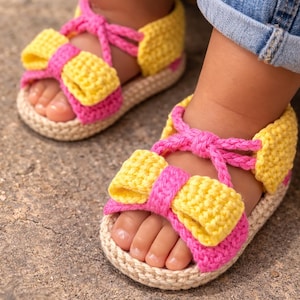 May include: Crocheted baby sandals in yellow, pink, and beige. The sandals feature a bow design on the front and a strap that ties around the ankle. The sandals are shown on a baby's feet, with blue jeans visible.
