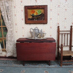 May include: Miniature dollhouse scene with a dark red table, a silver tea set on a tray, and a wooden chair. A framed painting of fruit hangs above the table. The room has floral wallpaper and a lace curtain.