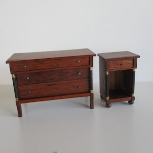 May include: Miniature wooden bedroom furniture set. The set includes a three-drawer dresser and a nightstand. The furniture is stained a dark reddish-brown with black and gold accents. The nightstand has a single drawer and an open shelf.