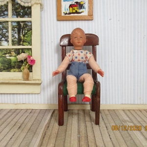 May include: A miniature doll with a tan complexion, short brown hair, and painted facial features, wearing a white shirt with a blue and red square pattern, blue shorts, and red shoes, seated on a dark brown wooden chair with a green seat.