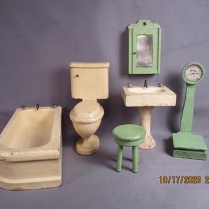 May include: A miniature bathroom set including a cream-colored bathtub, a matching toilet, a pedestal sink, a green medicine cabinet with a mirror, a small green stool, and a green standing scale with a dial labeled "scale" in black ink.
