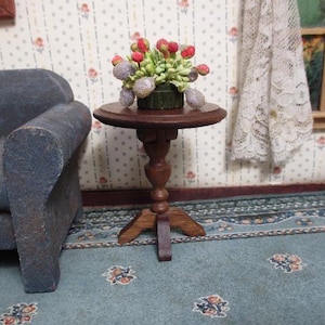 May include: A miniature round wooden pedestal table with a dark brown finish, featuring a central turned column and a tripod base. A small decorative arrangement of red and light purple floral buds with green foliage sits on the tabletop surface.