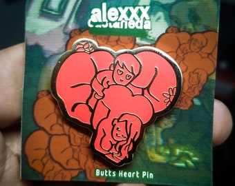 Puede incluir: Un pin rojo en forma de corazón con un contorno dorado, con figuras de dibujos animados. El pin está en una tarjeta con el texto "alexxx castaneda" y "Butts Heart Pin". El fondo tiene una ilustración de figuras.