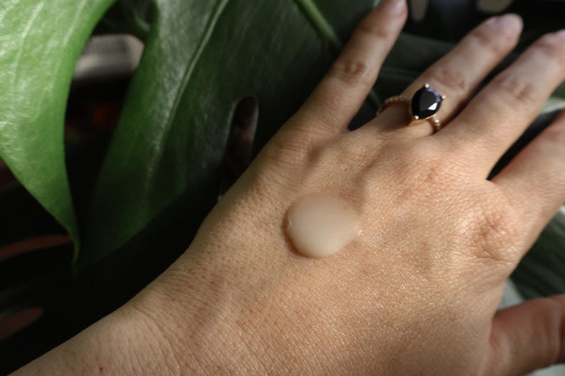 May include: A close-up of a hand with a clear, viscous liquid on the palm. The liquid is slightly pearlescent and has a smooth, even texture. The hand is wearing a gold ring with a black gemstone.