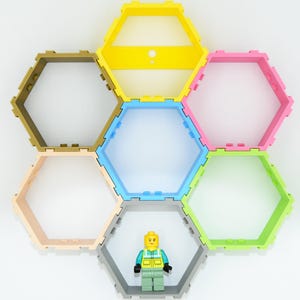 Op de afbeelding: Zes zeshoekige planken in geel, lichtblauw, roze, groen, beige en grijs, gerangschikt in een honingraatpatroon op een witte muur. Een klein geel en groen Lego-figuurtje staat in het midden van de planken.