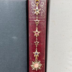 May include: A gold bookmark with a chain of stars and a sun with a pink centre. The bookmark is hanging from a book.