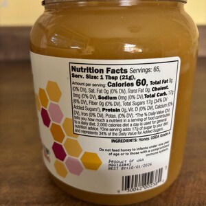 May include: A clear glass jar of honey with a white lid and a nutrition facts label. The label states that one tablespoon contains 60 calories and 17 grams of total carbohydrates. The label also includes a list of ingredients, which includes honey and USDA Grade A. The jar has a pink and yellow honeycomb design on the side.