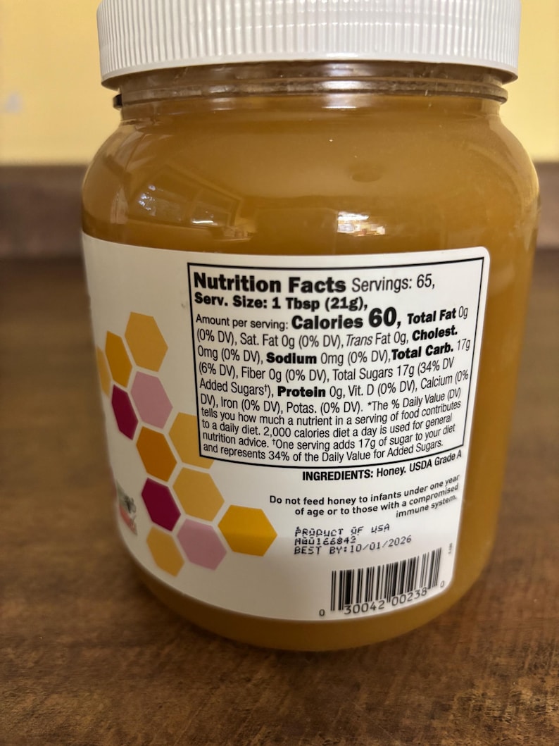 May include: A clear glass jar of honey with a white lid and a nutrition facts label. The label states that one tablespoon contains 60 calories and 17 grams of total carbohydrates. The label also includes a list of ingredients, which includes honey and USDA Grade A. The jar has a pink and yellow honeycomb design on the side.