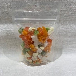 Puede incluir: Una bolsa de plástico transparente llena de una colorida mezcla de frutas deshidratadas y chispas de chocolate blanco. Las piezas de fruta son naranjas, verdes y blancas, con una variedad de formas y tamaños. La bolsa está sellada en la parte superior.