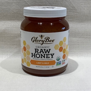May include: A glass jar of Glory Bee Organic Raw Honey, pure and unfiltered, light and mild. The jar is labeled with a bee and honeycomb design. The jar is 48 ounces (1.36 kg).