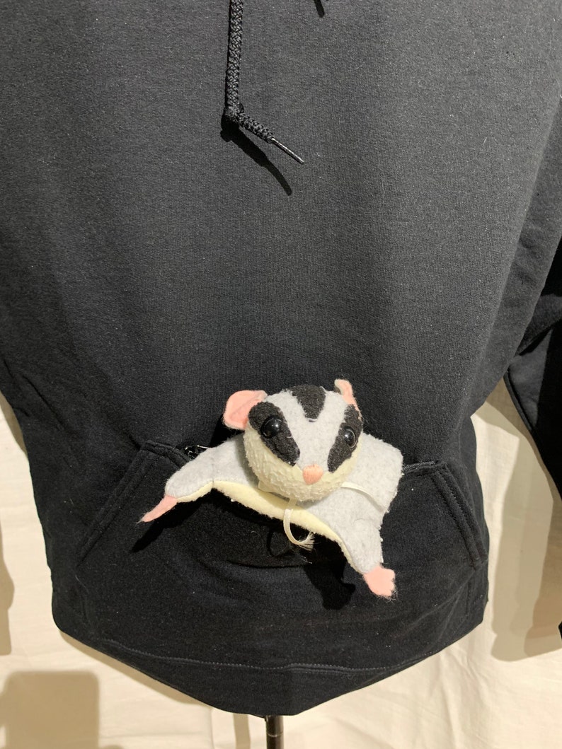 Op de afbeelding: Een zwarte hoodie met een grote zak aan de voorkant. Een klein wit en zwart knuffeldier zit in de zak.