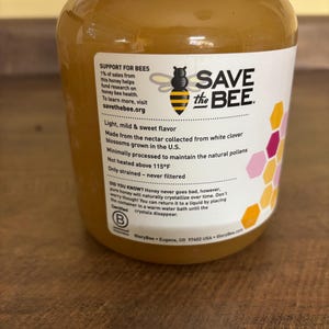 May include: A glass jar of honey with a white label that says "Save the Bee". The label also includes information about the honey, such as its flavor, origin, and how it is processed. The jar is sitting on a wooden surface.