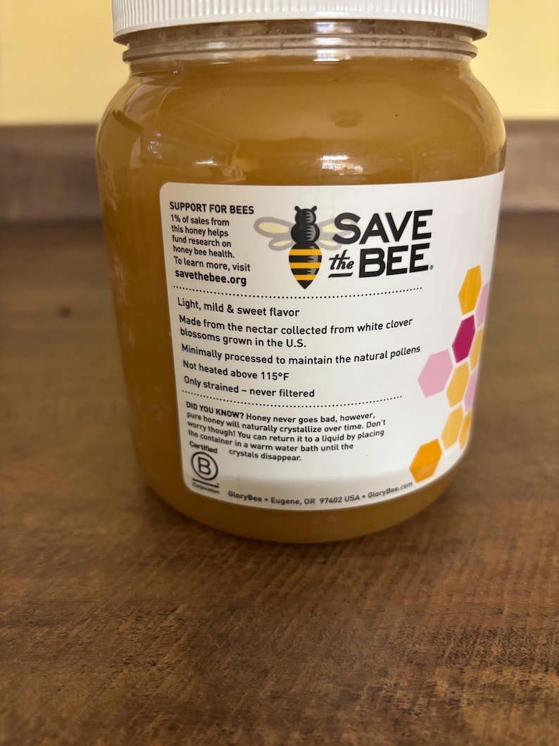 May include: A glass jar of honey with a white label that says "Save the Bee". The label also includes information about the honey, such as its flavor, origin, and how it is processed. The jar is sitting on a wooden surface.