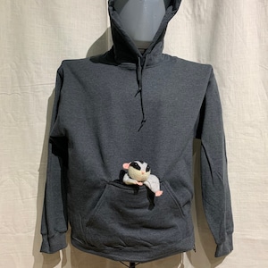 Op de afbeelding: Een donkergrijze hoodie met een grote zak aan de voorkant. Een klein wit en grijs knuffeldier zit in de zak.