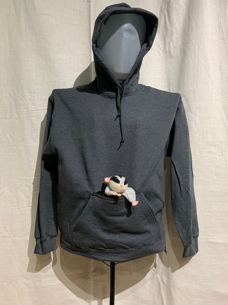 Op de afbeelding: Een donkergrijze hoodie met een grote zak aan de voorkant. Een klein wit en grijs knuffeldier zit in de zak.