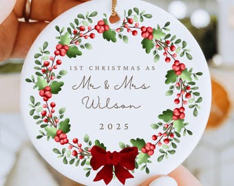 Puede incluir: Adorno navideño redondo de cerámica blanca con una corona de bayas rojas y hojas verdes. El texto dice "1st Christmas as Mr & Mrs Wilson 2025" con un lazo rojo. Incluye una cuerda dorada para colgar.