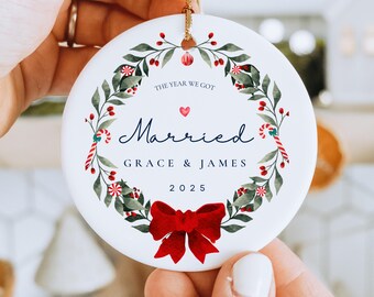 Puede incluir: Adorno navideño redondo de cerámica blanca con un diseño de corona. La corona incluye hojas verdes, bayas rojas y bastones de caramelo. El texto dice "The Year We Got Married", con nombres y el año "2025". Un lazo rojo adorna la parte inferior.