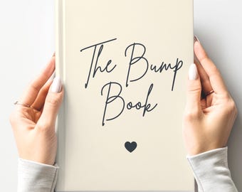 May include: A cream-colored book titled "The Bump Book" in elegant black script, with a small black heart beneath the title. The book is held by two hands with manicured nails, suggesting a journal or keepsake.