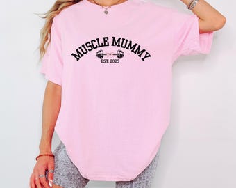 Puede incluir: Camiseta rosa de gran tamaño con el texto "Muscle Mummy Est. 2025" y un gráfico de una barra con pesas.
