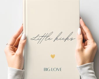 May include: A cream-colored journal with the words "Little kicks" in elegant script and a gold heart. The phrase "BIG LOVE" is printed below. The journal is held by a person.