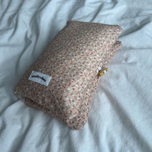 May include: A quilted rectangular pouch featuring a dusty pink fabric with a small-scale floral pattern in white, red, and green. The pouch has a white rectangular label with the black text studio viken and a small wooden toggle button closure.